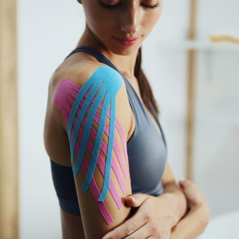 Sportive woman sits indoors with kinesio tape on her shoulder.